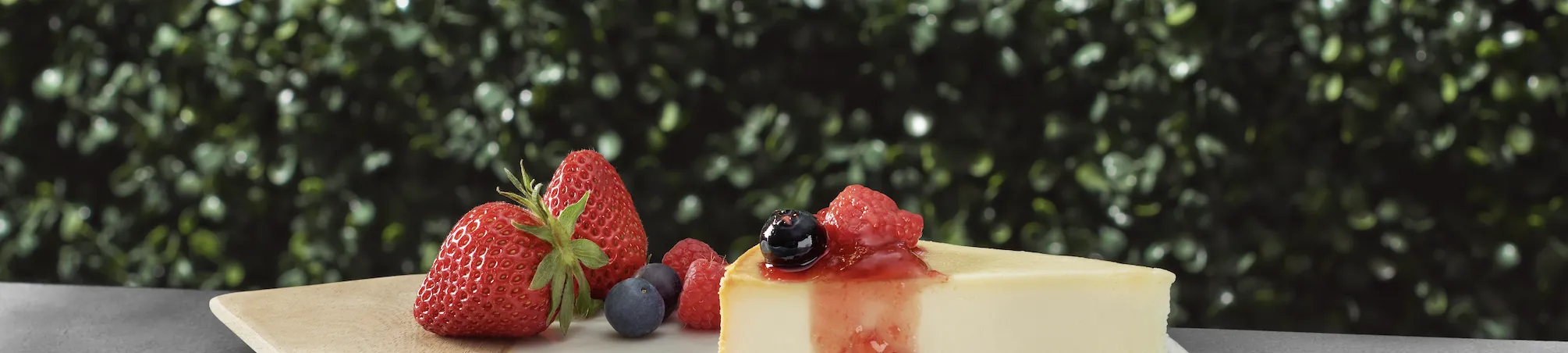 Cheesecake topped with berries, in front of a bush