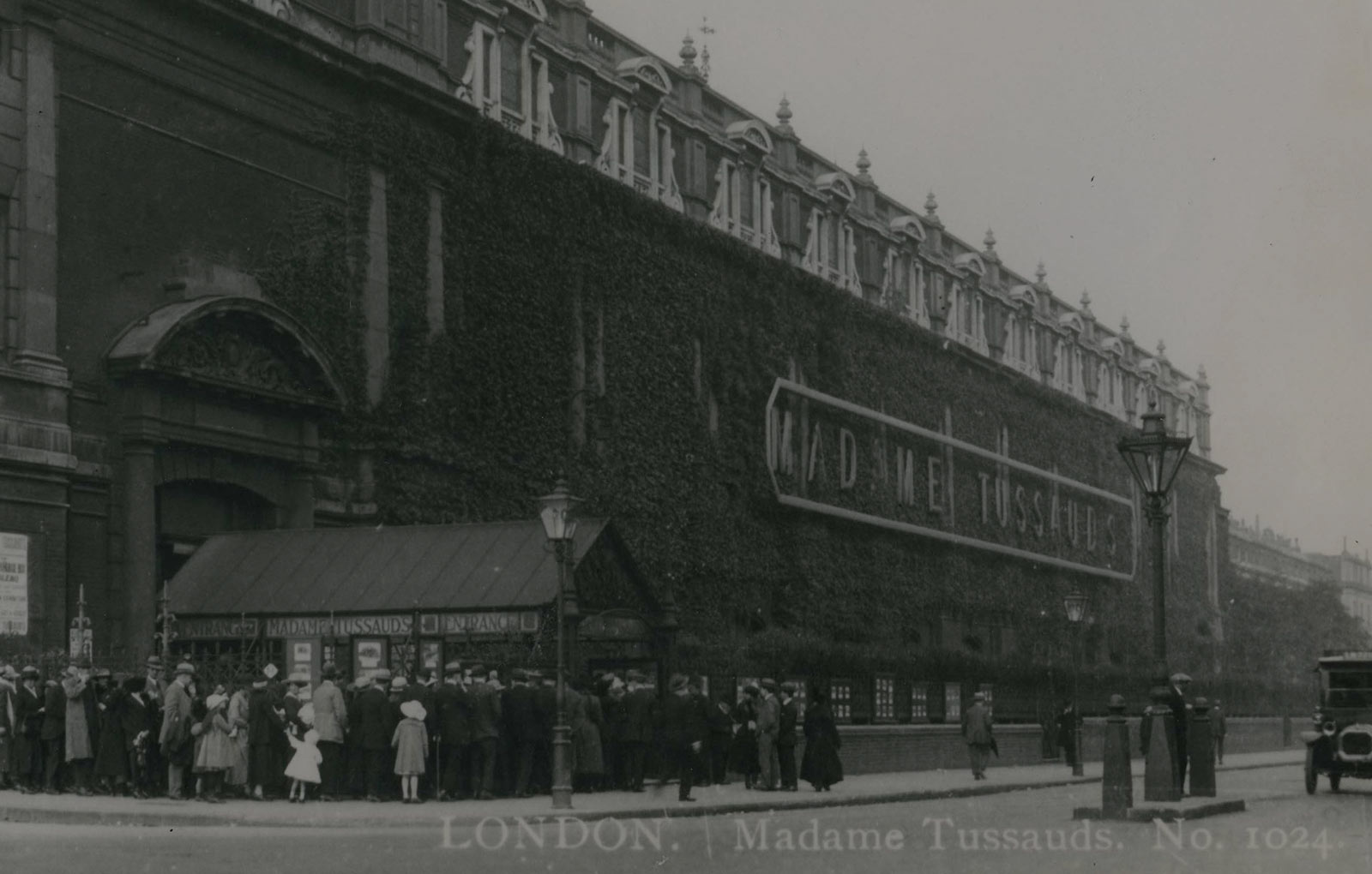 A historical black and white photograph of the exterior of Madame Tussauds London building. The building has a neoclassical facade with columns and ornate decorations, and a sign with the Madame Tussauds logo is visible above the entrance. 