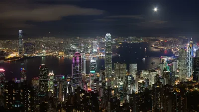 A panoramic view of Hong Kong at night, with the skyscrapers of  Central lit up against the dark sky. The Victoria Harbour is also visible, with the Star Ferry crossing in the foreground.