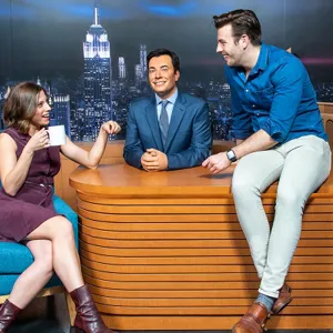One guest posing and another guest sitting on a desk with Jim Fallon's wax figure
