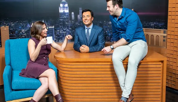 One guest posing and another guest sitting on a desk with Jim Fallon's wax figure