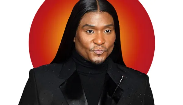 Portrait of celebrity, Law Roach with dark straight sleek hair, wearing a black turtleneck shirt under a black suit jacket outfit. A vibrant red and orange circular background highlights his face. Featured in Madame Tussauds' 'Hot 100' list.