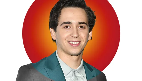 Portrait of celebrity, Marcello Andres Hernández Gonzales with dark wavy hair, wearing a light gray polo shirt under a dark gray suit jacket outfit. A vibrant red and orange circular background highlights his face. Featured in Madame Tussauds' 'Hot 100' list.
