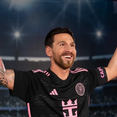 A wax figure of Lionel Messi at Madame Tussauds Orlando, dressed in an Inter Miami CF jersey, smiling with both arms raised triumphantly in a stadium setting illuminated by bright lights.