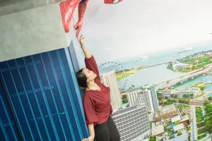 A visitor at Madame Tussauds Singapore - Marvel Universe 4D, pointing towards a Spider-Man figure that appears to be clinging to the side of a building exterior. The visitor is standing in front of a cityscape backdrop, making it seem like they are part of a thrilling superhero adventure