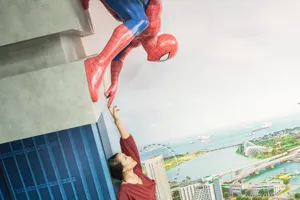 A visitor at Madame Tussauds Singapore - Marvel Universe 4D, pointing towards a Spider-Man figure that appears to be clinging to the side of a building exterior. The visitor is standing in front of a cityscape backdrop, making it seem like they are part of a thrilling superhero adventure