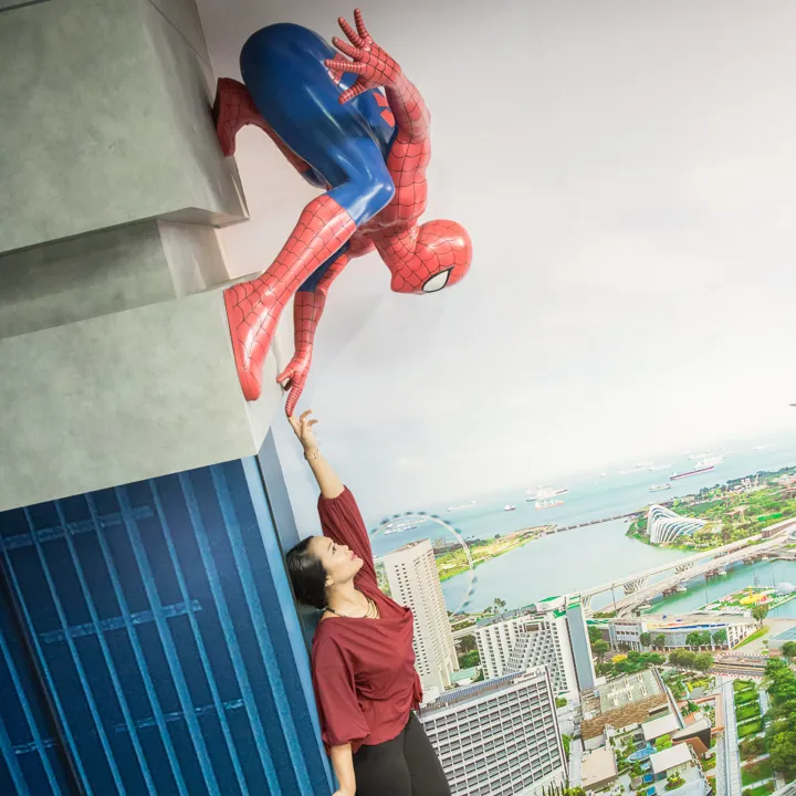 A visitor at Madame Tussauds Singapore - Marvel Universe 4D, pointing towards a Spider-Man figure that appears to be clinging to the side of a building exterior. The visitor is standing in front of a cityscape backdrop, making it seem like they are part of a thrilling superhero adventure