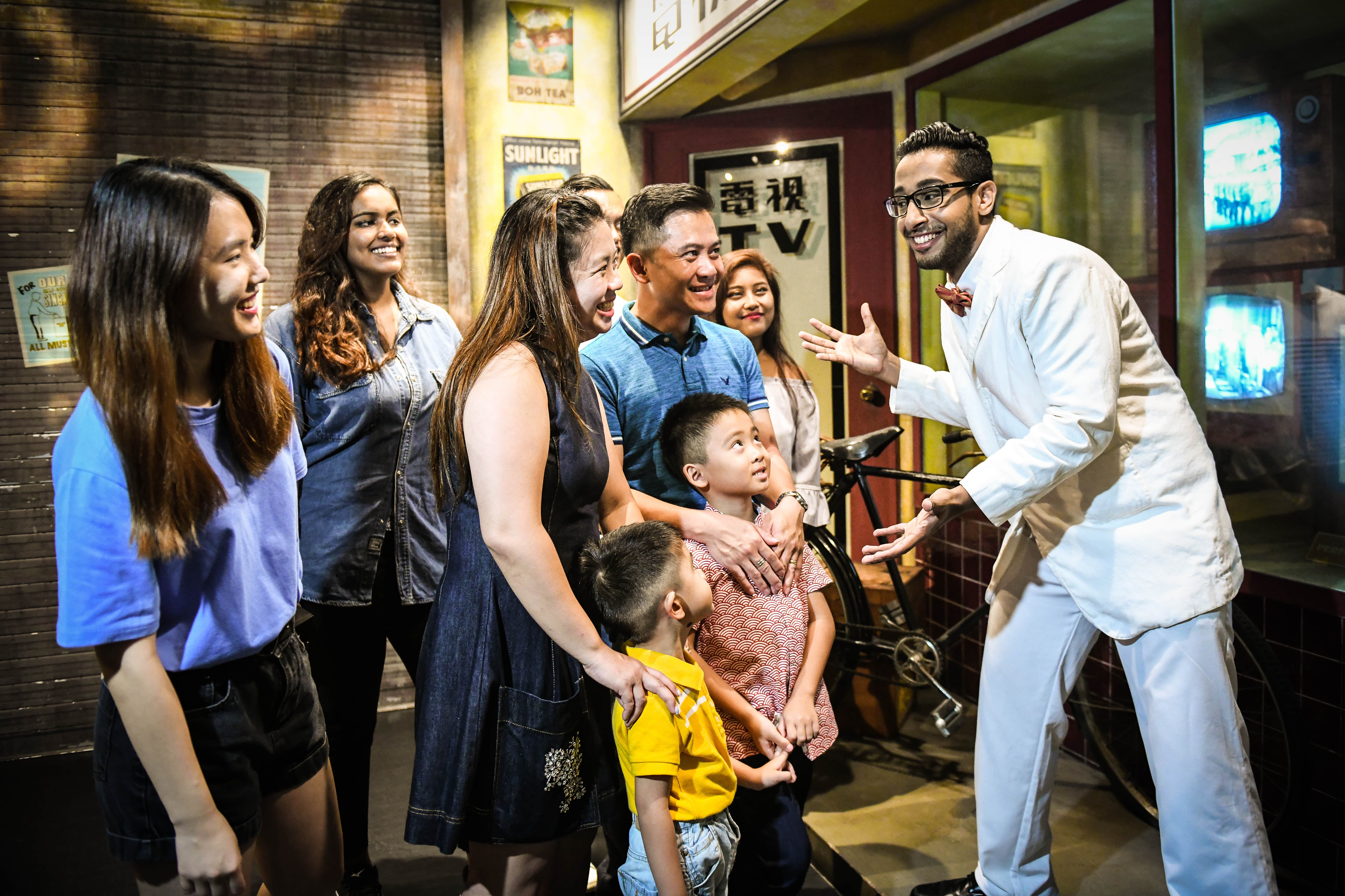 8 visitors including men, women and children in the exhibition of Images of Singapore, are beaming joyfully while looking at the wax figure salesman in a white suit outside the television shop smiling back at them with a welcoming gesture.