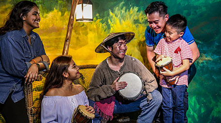 In the set-up of a Malay fishing village is a wax figure of a man in a straw hat. He smiles as he plays the drum, while three visitors of the Singapore exhibition joyfully look at him while acting to play the drum along. 