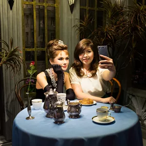 A woman taking a selfie with a wax figurine of Audrey Hepburn at Madame Tussauds Singapore TV & Film Zone. The wax figurine is dressed in a black gown with pearl earrings and necklace. 