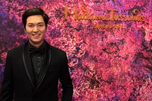 At Madame Tussauds Singapore, the wax figure of Korean celebrity and actor Lee Min Ho, dressed in a black suit and ribbon tie, smiles while standing against a backdrop of cherry blossoms