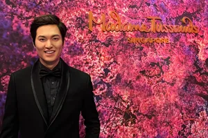 At Madame Tussauds Singapore, the wax figure of Korean celebrity and actor Lee Min Ho, dressed in a black suit and ribbon tie, smiles while standing against a backdrop of cherry blossoms